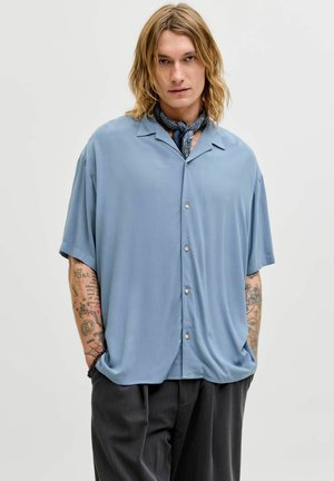 Jeune personne aux longs cheveux blonds portant une chemise bleu clair à manches courtes boutonnée, un foulard noir autour du cou et un pantalon noir, les mains dans les poches, fond neutre.