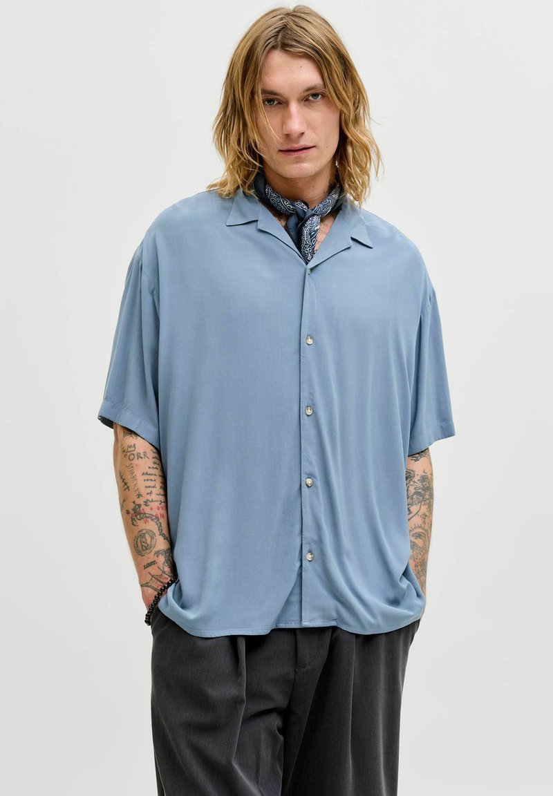 Młoda osoba o długich blond włosach nosi jasnoniebieską, krótkorękawową, zapinaną na guziki koszulę, czarną apaszkę na szyi oraz czarne spodnie, ma ręce w kieszeniach, neutralne tło.