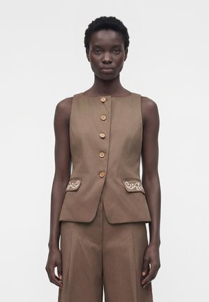 Slank kvinde iført en brun ærmeløs vest med knapper og blomsterbroderi på lommeklapperne, kombineret med matchende vidde bukser.