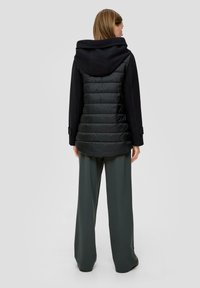 Doudoune noire avec capuche, présentant un design matelassé, associée à un pantalon taille haute à jambes larges en tissu vert foncé.