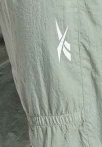 Lysegrønt, krøllet stof med elastisk manchet. Har en hvid geometrisk logo på siden. Glat tekstur med en afslappet pasform.