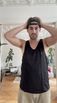 Camiseta sin mangas negra con logo en el pecho, usada por una persona, combinada con pantalones cortos de color claro en una habitación con plantas y muebles.