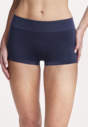 Marineblaue nahtlose Boyshort-Unterwäsche, getragen von einer stehenden Frau, die von der Taille bis zur Mitte des Oberschenkels vor einem einfarbigen Hintergrund zu sehen ist.