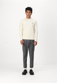 Cream cable-knit sweater with ribbed hem, paired with tailored gray trousers and black loafers. Simple, modern design with green logo accent.