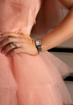 Montre en argent avec un cadran rectangulaire et des accents en cristal, accompagnée d'une bague. La main repose sur un tissu de tulle rose superposé d'une robe.