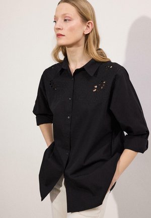 Γυναίκα με ξανθά μαλλιά που φοράει μαύρο πουκάμισο με κουμπιά και λουλουδένια σχέδια με ανοίγματα, με τα μανίκια σηκωμένα, τα χέρια στις τσέπες, να στέκεται μπροστά σε απλό φόντο.