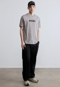 Γκρι T-shirt με στρογγυλή λαιμόκοψη και μαύρο λογότυπο "BOSS", φαρδιά μαύρη παντελόνια και μπεζ αθλητικά παπούτσια. Το μοντέλο φοράει μαύρο καπέλο με το γράμμα "BOSS".