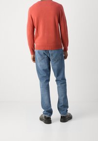 Homme portant un pull à manches longues corail, un jean bleu avec des poches arrière et des chaussures noires, se tenant contre un fond blanc uni.