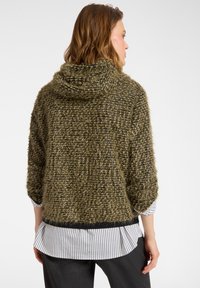 Olijf groene, gestructureerde gebreide hoodie met een losse pasvorm, gecombineerd met een wit en zwart gestreept shirt eronder en donkere broek.