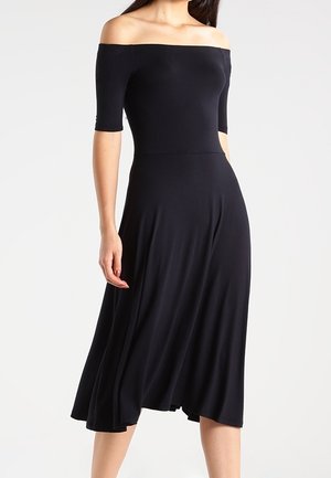 Frau trägt ein figurbetontes schwarzes Midi-Kleid mit schulterfreiem Ausschnitt, Ellbogenlangen Ärmeln und ausgestelltem Rock vor einem schlichten Hintergrund.