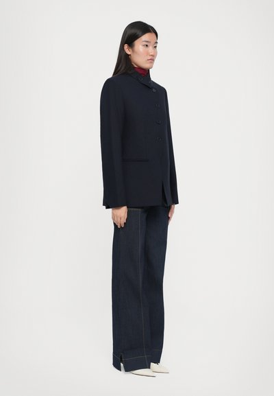 Marineblauwe wollen jas met vijf knopen, slim fit, en zijzakken, gecombineerd met hoog-gewijde donkere denim broek en puntige witte schoenen.