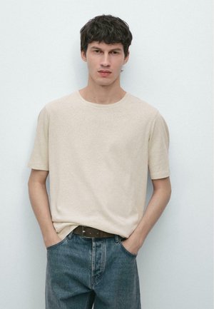 Giovane uomo con capelli corti e scuri in piedi contro una parete bianca, indossa una camicia beige a maniche corte e jeans blu con le mani nelle tasche.