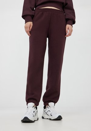 Pantaloni della tuta bordeaux con cintura e polsini elasticizzati, realizzati in tessuto morbido. Abbinati a sneaker sportive bianche e nere.