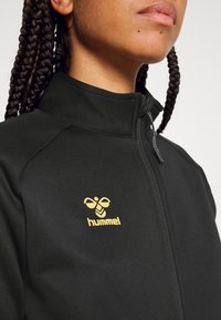 Gros plan d’une personne portant une veste noire zippée avec le logo jaune hummel, montrant des cheveux tressés et la partie inférieure du visage avec des piercings aux lèvres.