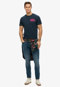 Camiseta de algodón azul marino con estampado gráfico en rosa, combinada con jeans azules desgastados y una camisa de franela a cuadros roja/verde atada en la cintura. Zapatillas ligeras.