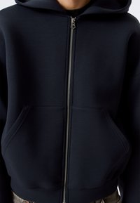 Sweat à capuche zippé bleu marine en tissu doux, avec une fermeture éclair à l'avant, deux grandes poches et une capuche doublée. Design minimaliste sans motifs visibles.