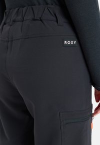 Czarne spodnie Roxy z elastycznym paskiem w talii, boczną kieszenią z zamkiem i minimalistycznym logo. Gładka tekstura materiału z dopasowanym krojem.
