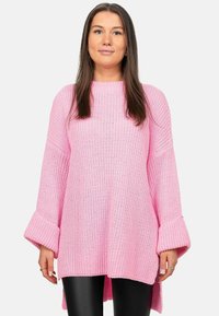 Roze gebreide trui met een losse pasvorm, geribbelde textuur, brede klokmouwen en een high-low onderlijn, gecombineerd met zwarte leggings.