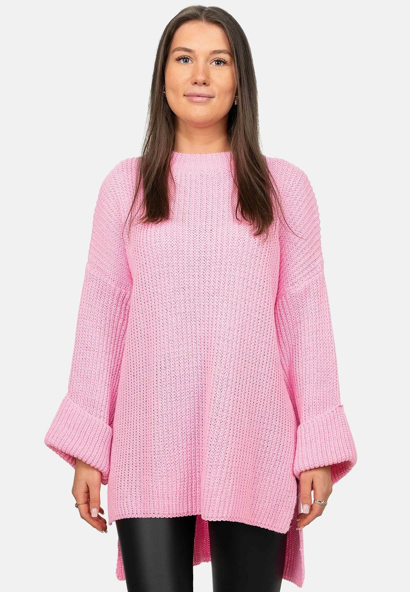 Roze gebreide trui met een losse pasvorm, geribbelde textuur, brede klokmouwen en een high-low onderlijn, gecombineerd met zwarte leggings.
