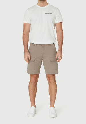 Mann trägt weißes kurzärmeliges T-Shirt mit kleinem Logo und beigefarbene Cargo-Shorts mit Klappentaschen, weiße Turnschuhe, steht vor einfarbigem Hintergrund.