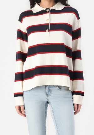 Kobieta ubrana w długi sweter z kołnierzykiem w poziome paski w kolorach granatowym, czerwonym i białym, w połączeniu ze spodniami jeansowymi w jasnoniebieskim kolorze.