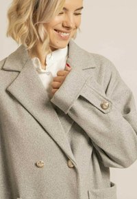 Manteau en laine gris clair avec un design croisé, grands revers, boutons dorés et tissu texturé. Comprend des poches latérales.