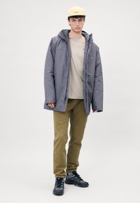 Veste matelassée grise avec capuche, chemise beige en dessous, pantalon vert olive et chaussures de sport noires. Design simple avec des accents minimalistes.