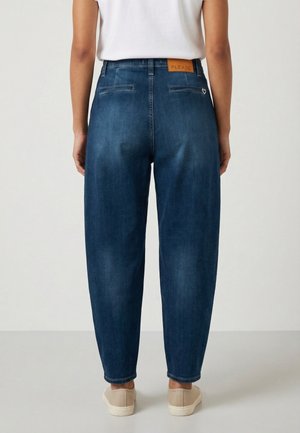 Persona che indossa jeans di denim blu scuro a vita alta e sneakers bianche, mostrata di schiena su uno sfondo chiaro e semplice.