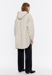 Chaqueta acolchada beige con capucha, con una silueta larga, un patrón texturizado en zigzag y puños elásticos, combinada con pantalones oscuros.