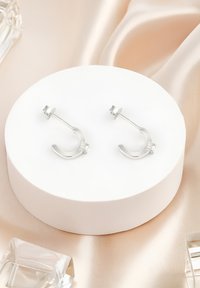 Silverfärgade hoop-örhängen med en slät, böjd design och en liten klar sten som accent. Monterade på ett runt vitt ställ.