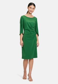 Grünes Kleid aus glattem Stoff, mit einem Bootsausschnitt, Dreiviertelärmeln und seitlicher Raffung für eine figurbetonte Silhouette.