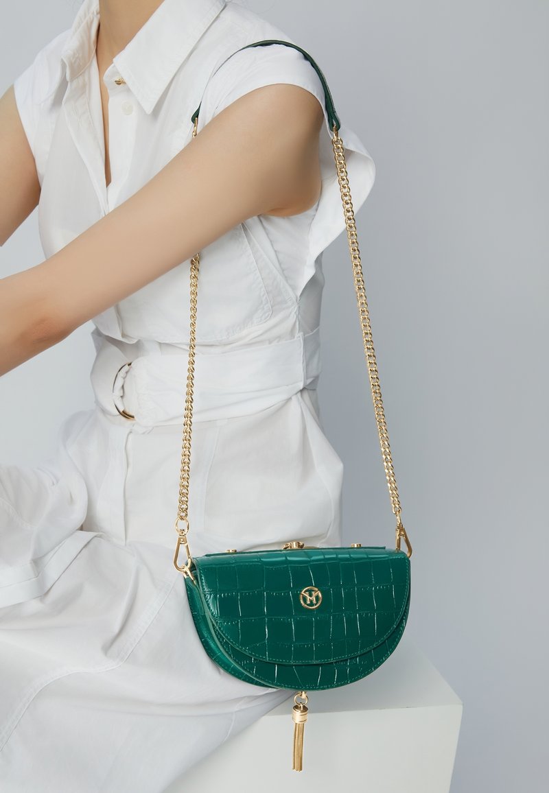 Femme en tenue blanche sans manches tient un sac à main vert à texture crocodile avec une bandoulière chaîne dorée et un détail pompon.