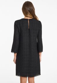 Zwarte tweed jurk met driekwart mouwen, een gestructureerd ruitpatroon en een franjezoom. Voorzien van een sleutelgat sluiting op de rug.