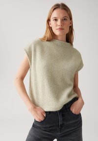Femme aux longs cheveux blonds portant un pull sans manches texturé vert clair et un jean noir, se tenant les mains dans les poches devant un fond uni.