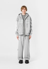 Sudadera gris con gráfico blanco en las mangas, combinada con pantalones de chándal grises a juego. El material parece suave; incluye cordones y bolsillos.