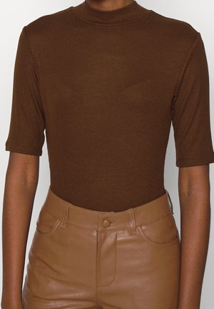 Personne portant un haut marron à manches courtes avec col montant, rentré dans un pantalon en cuir beige taille haute avec bouton et fermeture éclair.