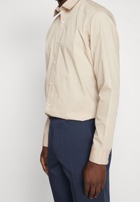 Beige Hemd mit spitzem Kragen, langen Ärmeln und weißen Knöpfen. Tragen zu dunkelblauen Hosen. Glatte Textur.