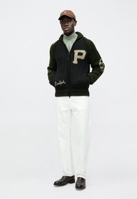 Camisola de malha preta e verde com fecho de correr e um grande "P" no peito, detalhes castanhos e bordado "Ralph". Usada com calças brancas e botas castanhas.