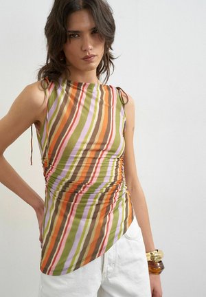 Femme portant un haut sans manches asymétrique rayé dans des tons orange, vert, marron et rose avec un pantalon blanc et des bracelets dorés et marron.