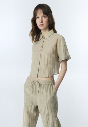 Jeune femme aux cheveux bruns portant une chemise beige texturée ample boutonnée et un pantalon assorti à cordon, mains dans les poches, fond neutre.