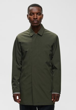 Homme portant un manteau long à manches longues vert olive, debout devant un fond gris clair uni.