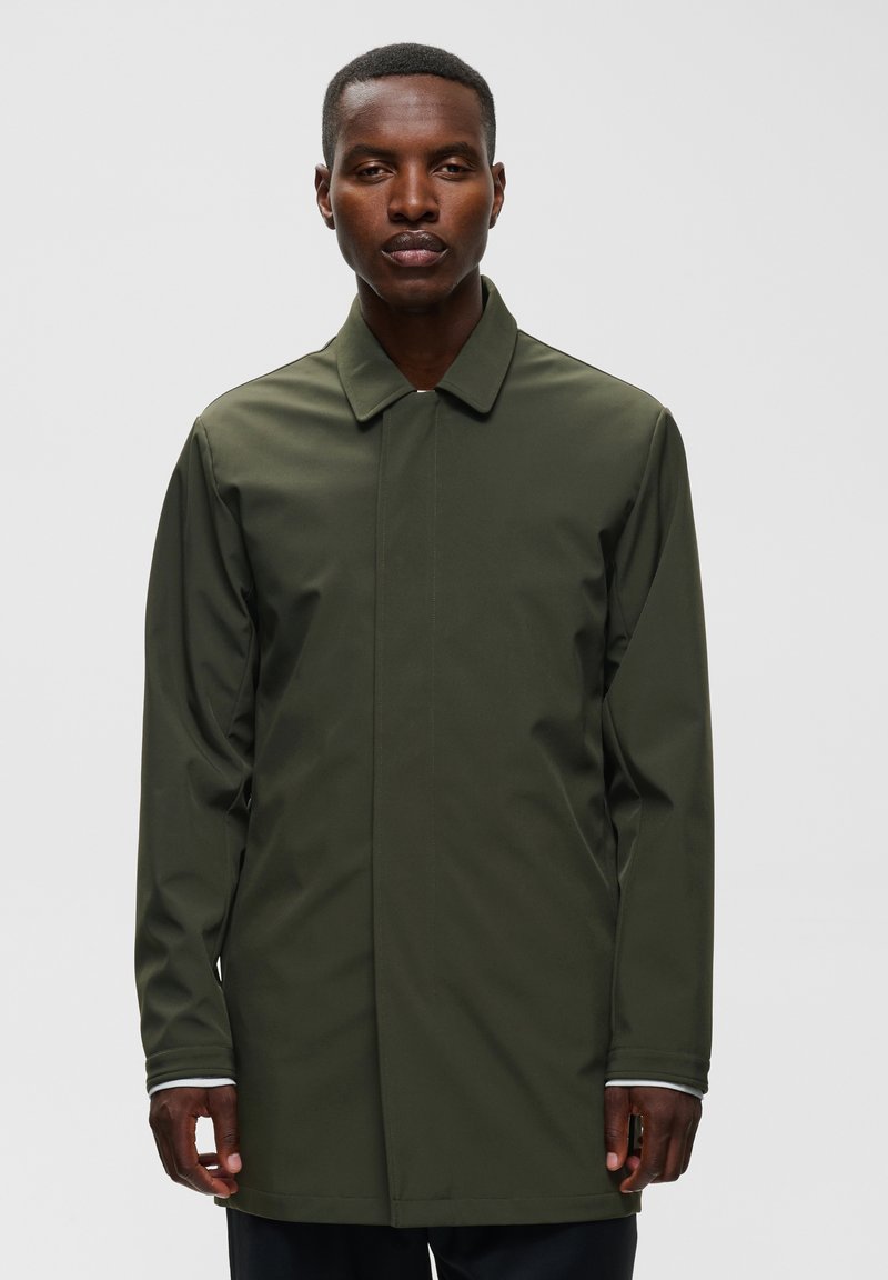 Homme portant un manteau long à manches longues vert olive, debout devant un fond gris clair uni.