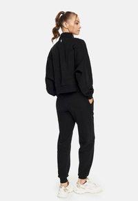 Schwarzes, kürzes Sweatshirt mit hohem Kragen und tailliertem Saum, kombiniert mit schwarzen, schmalen Jogginghosen. Beide bestehen aus weichem, strukturiertem Stoff.