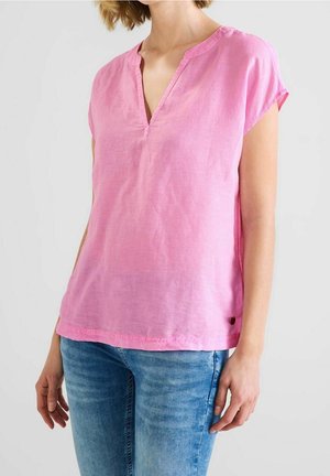 Frau trägt eine lockere, pinke Bluse mit kurzen Ärmeln und V-Ausschnitt sowie hellblaue Jeans vor einem einfarbigen Hintergrund.