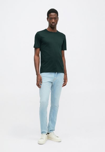 Σκούρο πράσινο t-shirt με κοντά μανίκια, συνδυασμένο με ανοιχτό μπλε τζιν και λευκά αθλητικά παπούτσια. Μοντέλο που στέκεται μπροστά από μια απλή, μονόχρωμη φόντο.