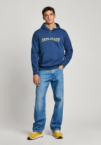 Navy blauwe hoodie met het gele "Pepe Jeans" logo, gecombineerd met lichtblauwe jeans en gele sneakers. Casual, relaxte pasvorm.