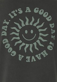 Camiseta de algodón negra con un gráfico central de un sol sonriente con rayos ondulados y el texto "ES UN BUEN DÍA PARA TENER UN BUEN DÍA" en verde claro.