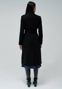 Zwarte wollen jas met een ceintuur in de taille, een gekartelde kraag en lange mouwen. Het model draagt het over een spijkerrok en zwart kniehoge laarzen.