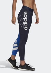 Osoba nosząca granatowe i niebieskie legginsy Adidas z dużym białym logo po boku oraz czarne buty do biegania z białymi podeszwami.