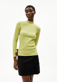 Langsleeve, geribbelde, limoengroene top met een hoge halslijn. Gecombineerd met een zwarte corduroy minirok en een label aan de zoom.
